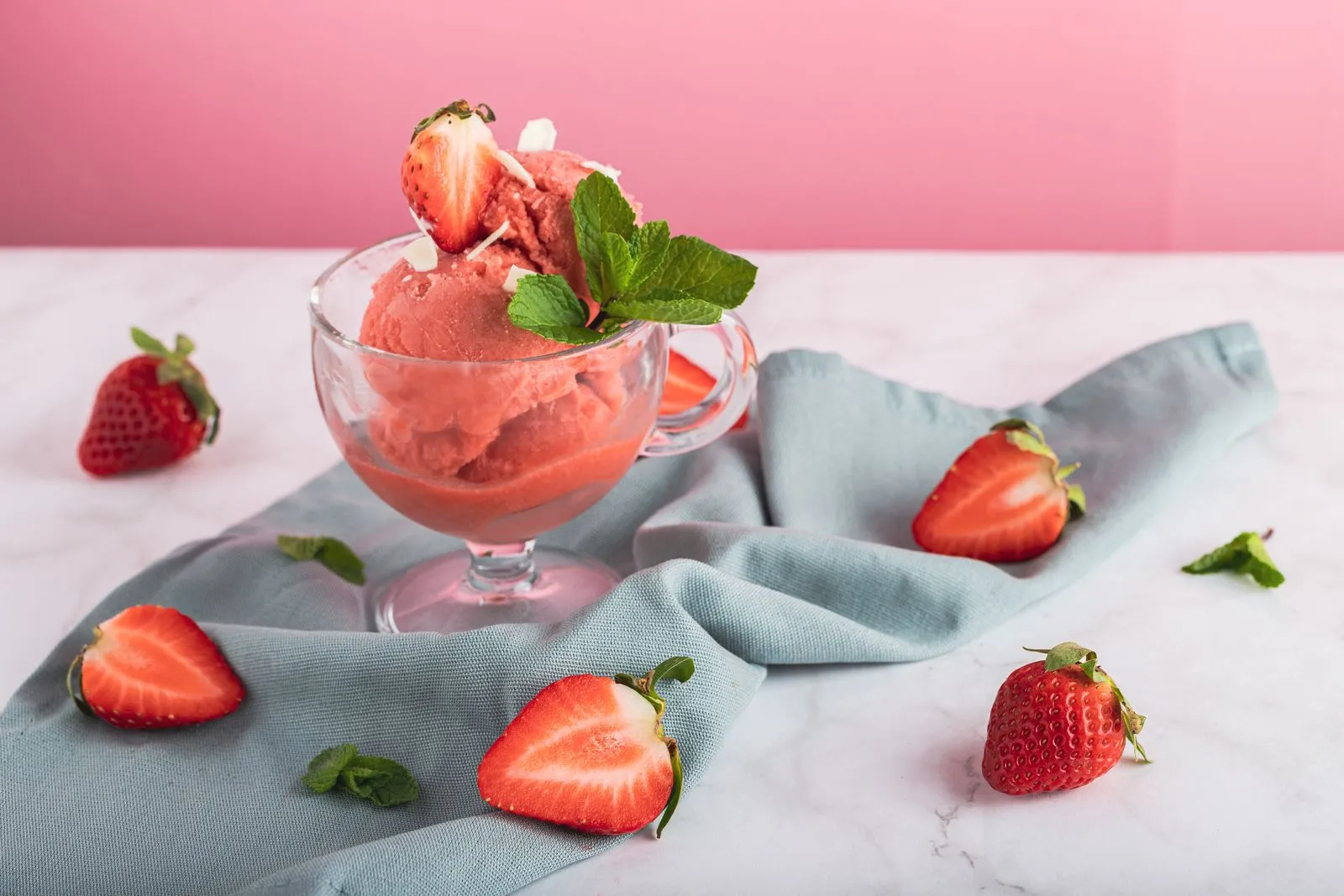 Refreshing strawberry sorbet in a glass, garnished with mint and fresh sliced strawberries on a pink background.